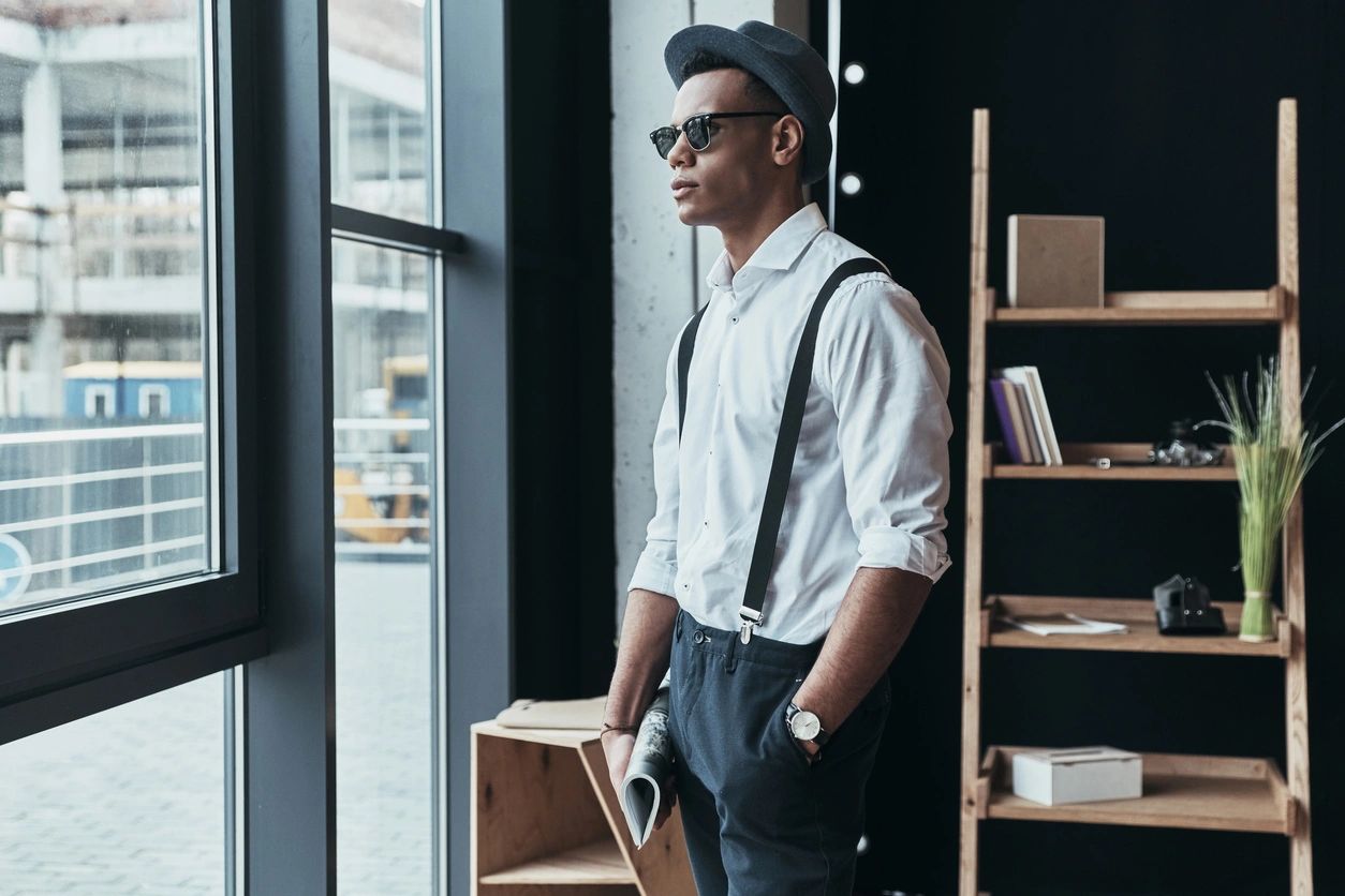 A stylish man in suspenders and a hat looks out a window, holding rolled-up blueprints in a modern office.