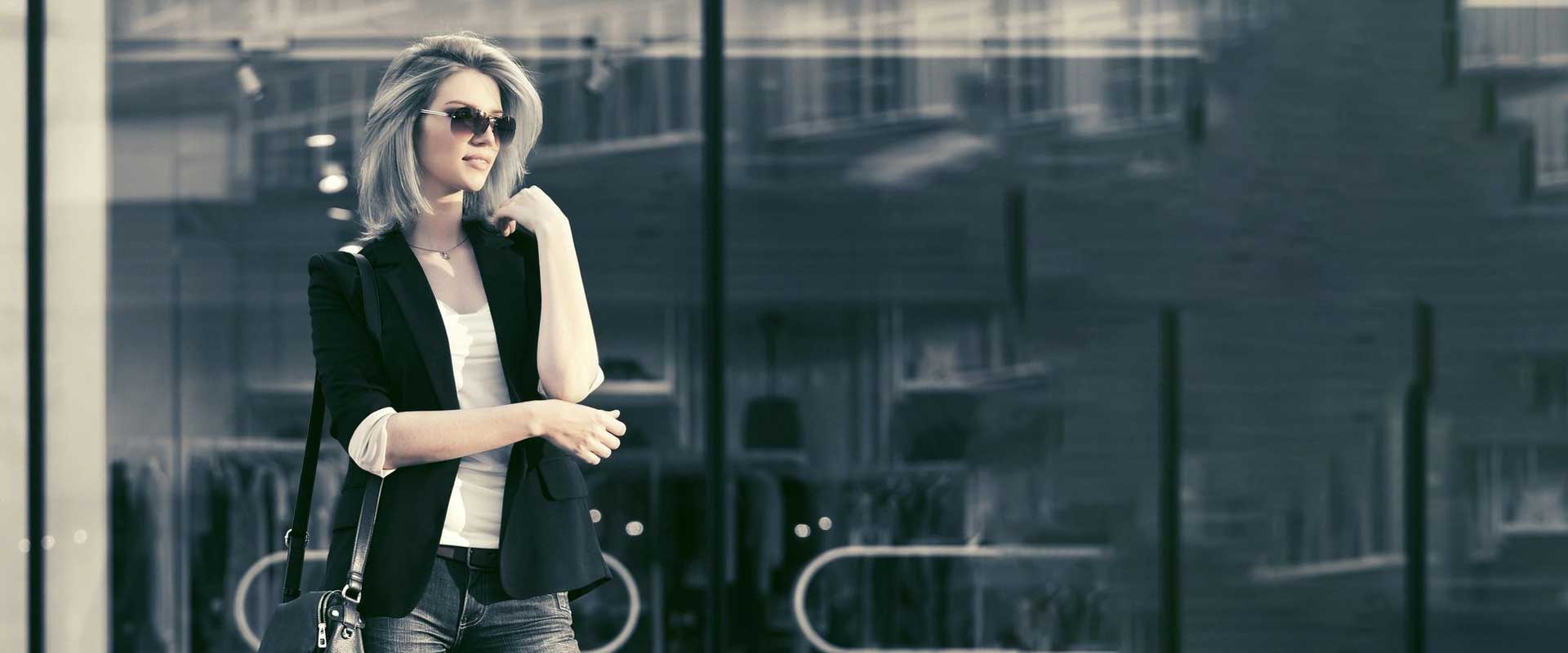 A stylish woman wearing sunglasses and a blazer, walking by a glass building in an urban setting.